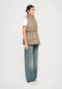 Beige gewatteerde vest met hoge kraag, een getailleerde taille en twee voorzakken, gecombineerd met lichtblauwe wijde jeans en puntige schoenen.