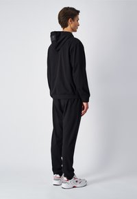 Outfit composto da felpa nera con cappuccio e pantaloni sportivi con fasce elastiche, realizzato in tessuto morbido. Caratteristiche notevoli includono un logo sul cappuccio e sneaker bianche.