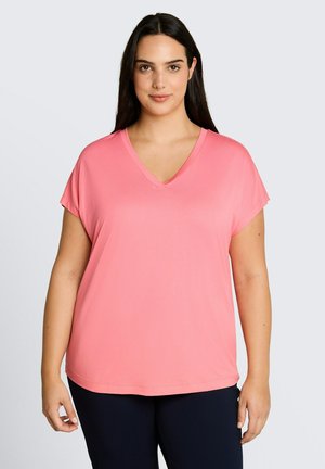 Donna con lunghi capelli scuri che indossa una maglietta rosa a maniche corte con scollo a V e pantaloni scuri, in piedi davanti a uno sfondo chiaro e uniforme.