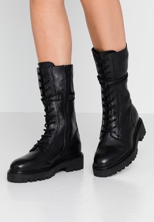 Bottes en cuir texturé noir mi-mollet avec lacets à l'avant, fermetures éclair latérales, semelles épaisses et robustes, portées sur des jambes nues contre un fond uni.