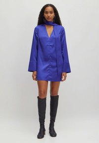 Satijnen mini-jurk in levendig blauw, met een V-hals en lange klokmouwen. Inclusief een bijpassende nekstrikdetail. Draag met zwartkniehoge laarzen.