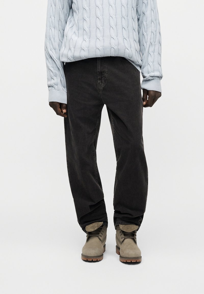 Personne portant un pull en maille torsadée bleu clair, un pantalon noir ample et des bottes en suède gris, debout sur un fond blanc.