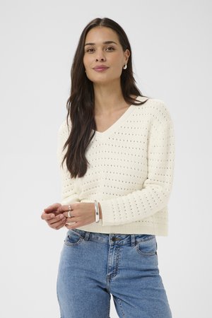 Frau mit langen dunklen Haaren, die einen weißen Strickpullover mit kleinen Löchern, blaue Jeans, ein silbernes Armband und Ringe trägt, steht vor einem schlichten Hintergrund.