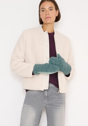 Ljusbeige shearlingjacka, lila stickad tröja, grå jeans och turkosa fleecevantar. Vantarna har en mjuk, strukturerad yta.