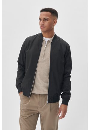 Mustade bomber-jakk, millel on sile materjal, lukuga sulgemine ja ribaline krae. Kanti beeži ülikonna üle ja kombineeritud heledate pükstega.