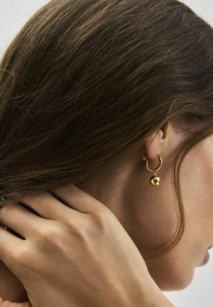 Frau mit braunen Haaren, die einen kleinen goldenen Creolenohrring mit einem runden, hängenden Anhänger trägt, Hand am Nacken liegend.