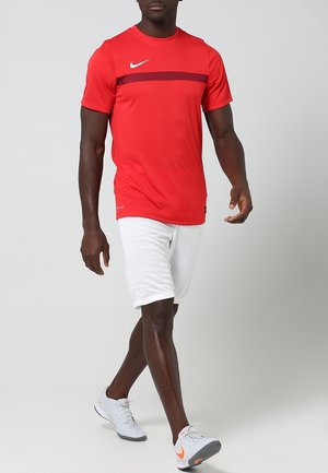 Maglietta da atletica rossa Nike con una striscia orizzontale color amaranto, abbinata a pantaloncini bianchi e scarpe da atletica grigie con dettagli arancioni.