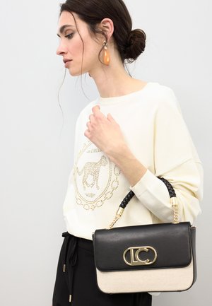 Zwarte en beige handtas met een gouden logo-accent en gevlochten handvat, gemaakt van leer en canvas. Model draagt een ivoren trui.