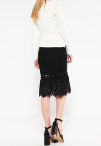 Schwarzer Spitzrock mit einem ausgestellten Saum, kombiniert mit einem gerippten weißen Oberteil. Das Outfit wird von schwarzen High Heels und einer glatten Textur ergänzt.