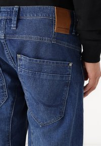 Blue denim jeans feature a leather patch labeled "JACK & JONES," large back pockets, and visible stitching detail.
