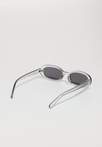 Gafas de sol ovaladas con montura transparente y lentes oscuros. Brazos delgados y rectos se extienden desde cada lado, mostrando un diseño minimalista.