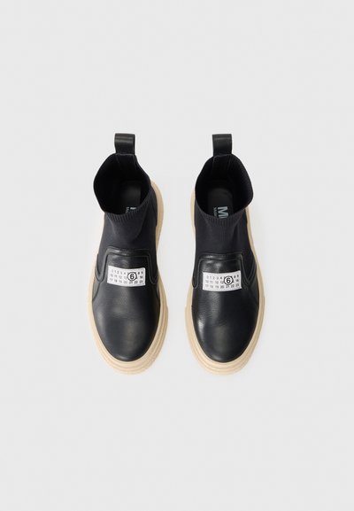 Svarta slip-on skor i läder och elastiskt material, med en vit etikett och sula i naturfärgad gummi.