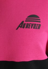 Pull-over fuchsia avec une bande noire en bas, arborant un logo imprimé en noir comprenant des rayures horizontales et du texte.
