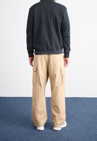Top negro de manga larga con canalé combinado con pantalones cargo beige holgados que tienen bolsillos laterales, llevado con zapatillas blancas sobre una alfombra azul.