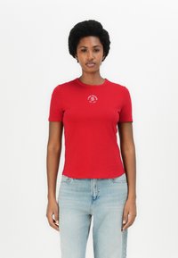 T-shirt en coton rouge à manches courtes, col rond, arborant un petit logo blanc sur la poitrine. Associé à un jean en denim bleu clair.