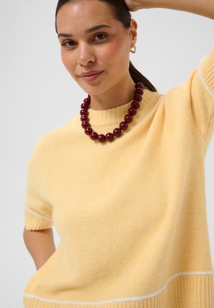 Femme aux cheveux foncés attachés en queue de cheval, portant un pull jaune à manches courtes et un collier de grosses perles rouge foncé, touchant sa tête.