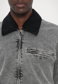 Veste zippée grise avec un col en velours côtelé noir. Dotée d'une poche poitrine avec une fermeture éclair. Logo texte "Armani Exchange Milano" sur le devant.