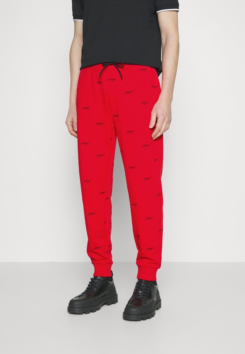 Rote Jogginghose mit Kordelzug am Bund, versehen mit einem wiederholten schwarzen Logo-Muster. Kombiniert mit schwarzen Schuhen mit klobiger Sohle. Glatte Textur.