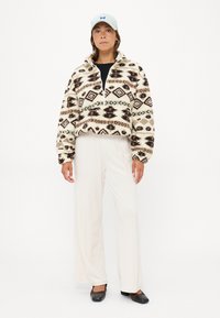 Fleece jakna z geometrijskimi vzorci v krem, rjavi in zeleni barvi, z visokim ovratnikom in sprednjim zadrgo. V kombinaciji s svetlimi hlačami in balerinkami.
