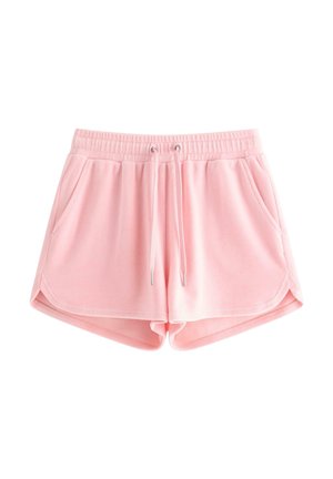 Ung pige med langt blondt hår iført en pink blomstret tanktop, lyserøde shorts med lommer og hvide sneakers, der står med hænderne i lommerne.
