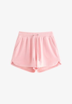 Ung pige med langt blondt hår iført en pink blomstret tanktop, lyserøde shorts med lommer og hvide sneakers, der står med hænderne i lommerne.
