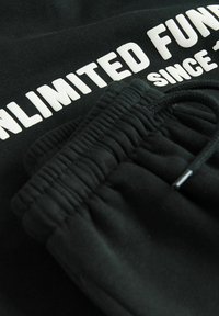 Schwarze Sweatpants mit elastischem Bund auf schwarzem Stoff, mit teilweise sichtbarem weißem Text "UNLIMITED FUN SINCE".