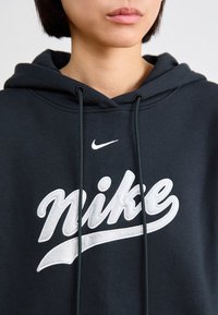 Närbild på en person som bär en svart huvtröja med vit Nike-logotyp och dragsko, med fokus på övre bröstet och läpparna.
