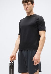 Svart sportt-shirt med kort ärm och rund halsringning, kombinerad med mörka shorts. En svart vattenflaska hålls i modellens högra hand.