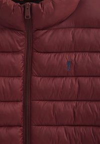 Burgundy vadderad väst med framsidig dragkedja, som har horisontella quiltade sektioner och en liten blå logotyp broderad på bröstet.