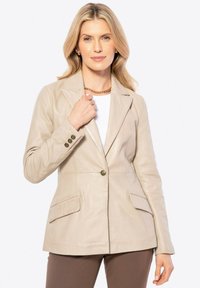 Blonde vrouw in een beige leren blazer boven een witte blouse en bruine broek, die met één hand de kraag van de blazer vasthoudt, neutrale achtergrond.