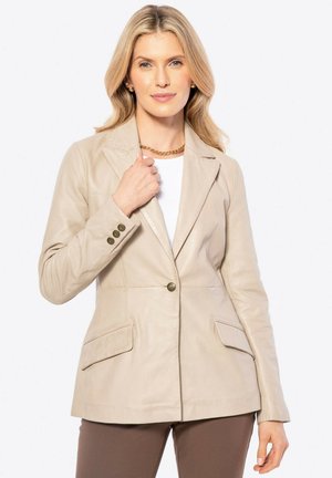 Blonde vrouw in een beige leren blazer boven een witte blouse en bruine broek, die met één hand de kraag van de blazer vasthoudt, neutrale achtergrond.
