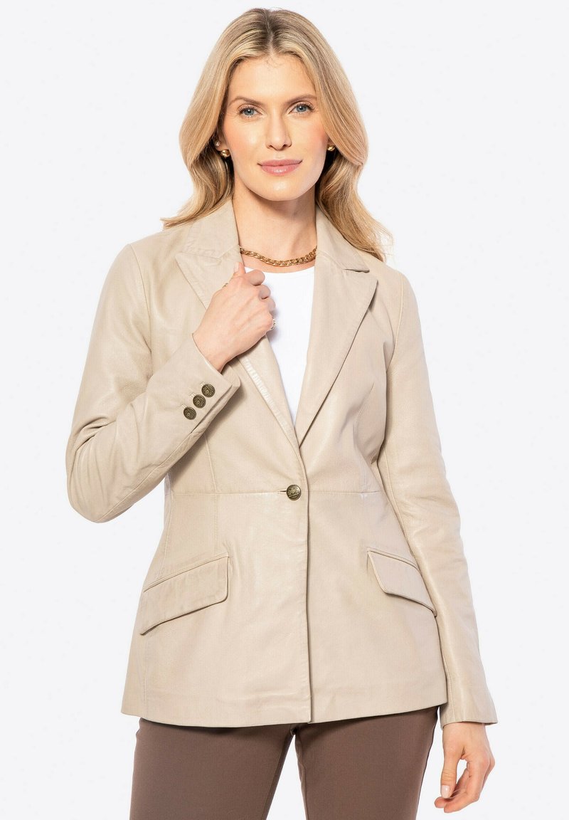 Blonde vrouw in een beige leren blazer boven een witte blouse en bruine broek, die met één hand de kraag van de blazer vasthoudt, neutrale achtergrond.