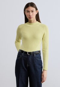 Κίτρινο πουλόβερ με ραβδώσεις, ψηλό γιακά και εφαρμοστή σχεδίαση, σε συνδυασμό με σκούρο τζιν και μαύρη ζώνη με λογότυπο.