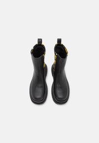 Pareja de botas de tobillo de cuero negro con paneles de tela floral amarilla en la parte posterior, lengüetas negras y suelas de goma gruesas.