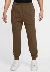 Pantaloni della tuta in cotone marrone con cintura in vita regolabile e polsini a costine, con un piccolo logo bianco sul lato sinistro, abbinati a sneakers bianche.