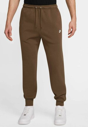 Brown cotton sweatpants with a drawstring waistband and ribbed cuffs, featuring a small white logo on the left side, paired with white sneakers.
