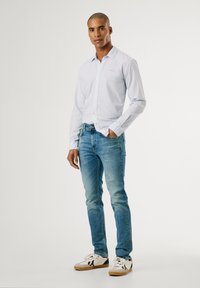 Camisa de rayas azul claro de manga larga, jeans ajustados de mezclilla y zapatillas blancas casuales con detalles en negro. Texturas suaves, diseño entallado.