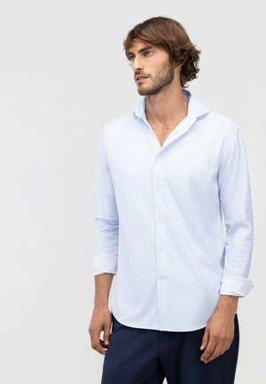 Hombre con cabello castaño claro y barba, vestido con una camisa de botones a rayas azul claro con las mangas remangadas y pantalones oscuros, mirando hacia la derecha.