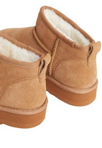 Des bottines en suède tan avec une douce doublure en fourrure blanche, un petit talon et des languettes à l'arrière pour un enfilage facile. Détails cousus le long des coutures.