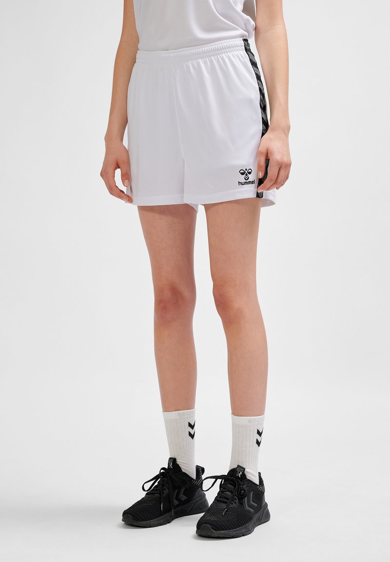 Person iført hvide sportsshorts med sorte striber og logo, hvide sokker med sorte chevronmønstre og sorte løbesko, stående på ensfarvet baggrund.