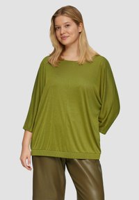 Vrouw met schouderlang blond haar, gekleed in een oversized olijfgroene top en donkere olijfgroene leren broeken, staand tegen een effen achtergrond.