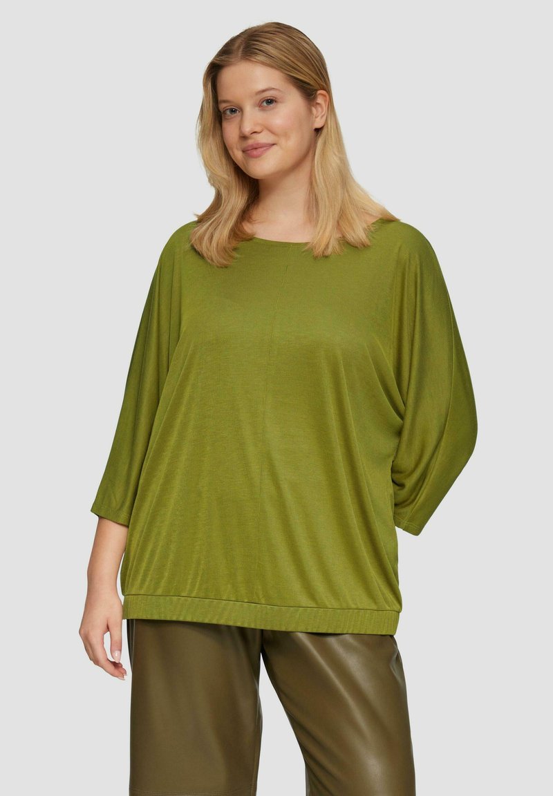 Vrouw met schouderlang blond haar, gekleed in een oversized olijfgroene top en donkere olijfgroene leren broeken, staand tegen een effen achtergrond.
