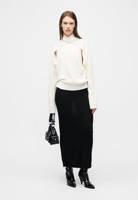 Femeie purtând un pulover alb cu guler înalt și decupaje, fustă midi neagră, cizme negre până la gleznă și ținând o geantă neagră.