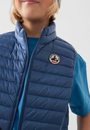 Enfant portant une veste sans manches matelassée bleue avec une fermeture éclair frontale et un écusson circulaire du logo JOTT, sur un t-shirt bleu.
