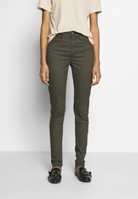 Vaqueros ajustados de color verde oliva, hechos de algodón, con cinco bolsillos, trabillas para el cinturón y cremallera. Combinados con una camiseta beige y mocasines negros.