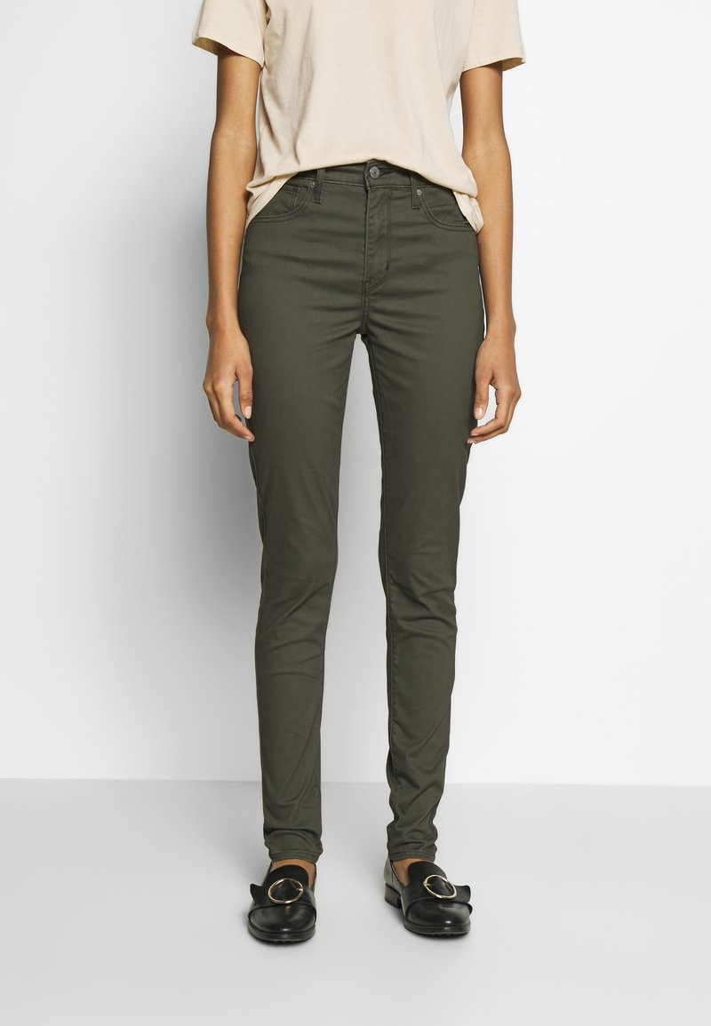 Vaqueros ajustados de color verde oliva, hechos de algodón, con cinco bolsillos, trabillas para el cinturón y cremallera. Combinados con una camiseta beige y mocasines negros.