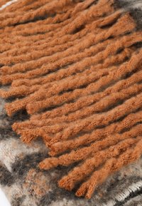 Textureret plaid med snoede, bløde orange frynser. Bunden har grå og kremfarvede nuancer med sorte accenter. Materialet ser varmt og fyldigt ud.