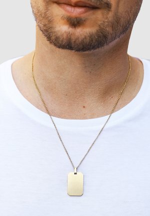 Eine goldfarbene Metallkette mit einem gravierten Hundemarkenanhänger mit abgerundeten Kanten, präsentiert vor einem schlichten weißen Hemd.