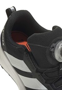 Μαύρο αθλητικό παπούτσι με άνω μέρος από δίχτυ, λευκές και γκρίζες λεπτομέρειες, κυκλικό κλείδωμα κορδονιών στην πλευρά και πορτοκαλί εσωτερική σόλα. Λεπτομέρειες με υφή στην επιφάνεια.