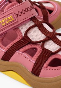 Gros plan d'une sandale d'enfant rose et marron en tissu mesh, bride réglable étiquetée "Banditos by Crio's" et semelle teintée jaune.